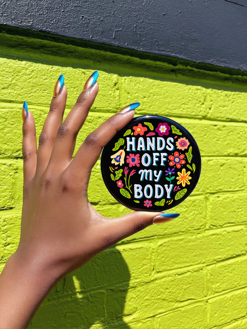 Glossy black pinback button with floral art and bold white text that reads “Hands Off My Body.” Feminist political button featuring bright pastel flowers in pink, red, orange, and yellow. Metal pin with durable safety pin back, shown in various settings: held by hand, displayed on pedestal, and against colorful street art. Available in 2" and 3" sizes, perfect for backpacks, jackets, tote bags, or as a gift for feminist allies and book lovers.